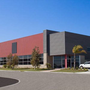 Office building, red and gray in color. This office building is facing the street and appears to be two stories. There are trees, a mailbox and glass front windows on this office building.